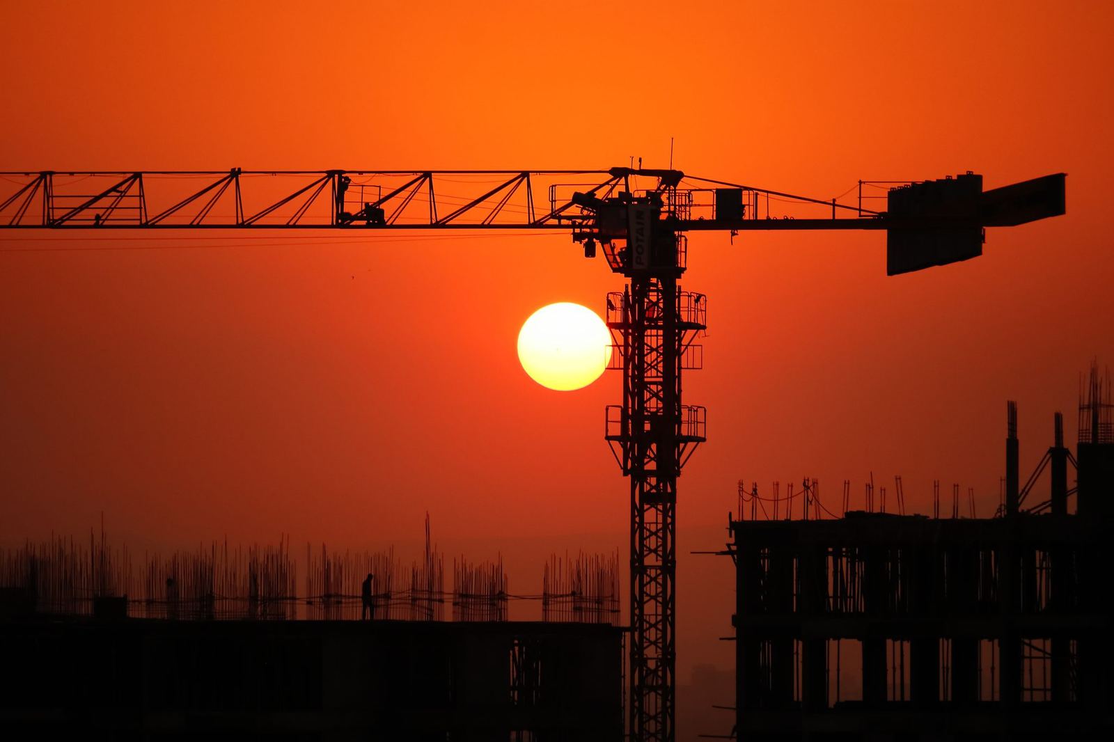 Silhouette of a tower crane and a building under construction against an orange sunset sky