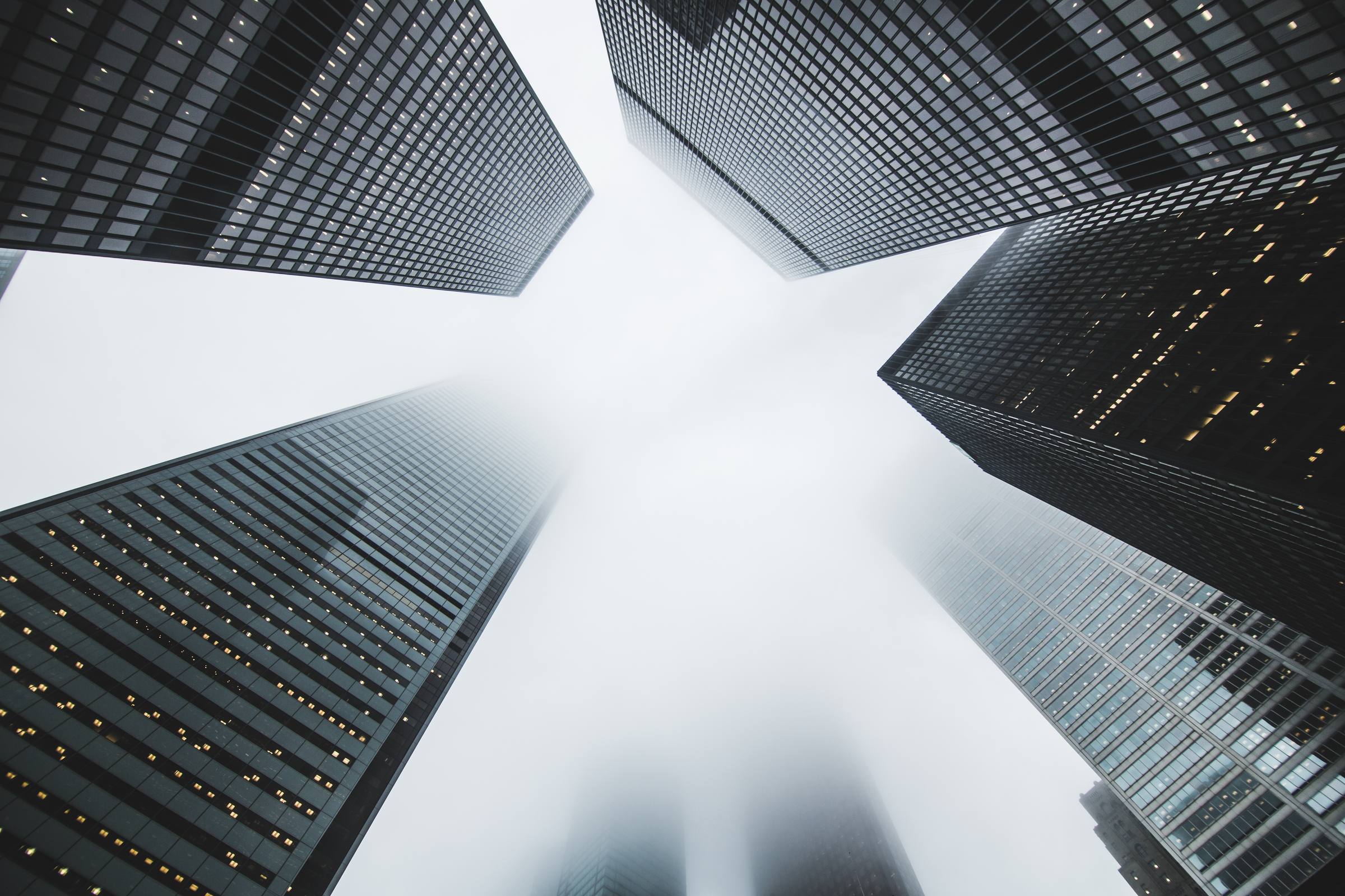Looking straight up between dark glass office towers whose tops disappear into thick fog