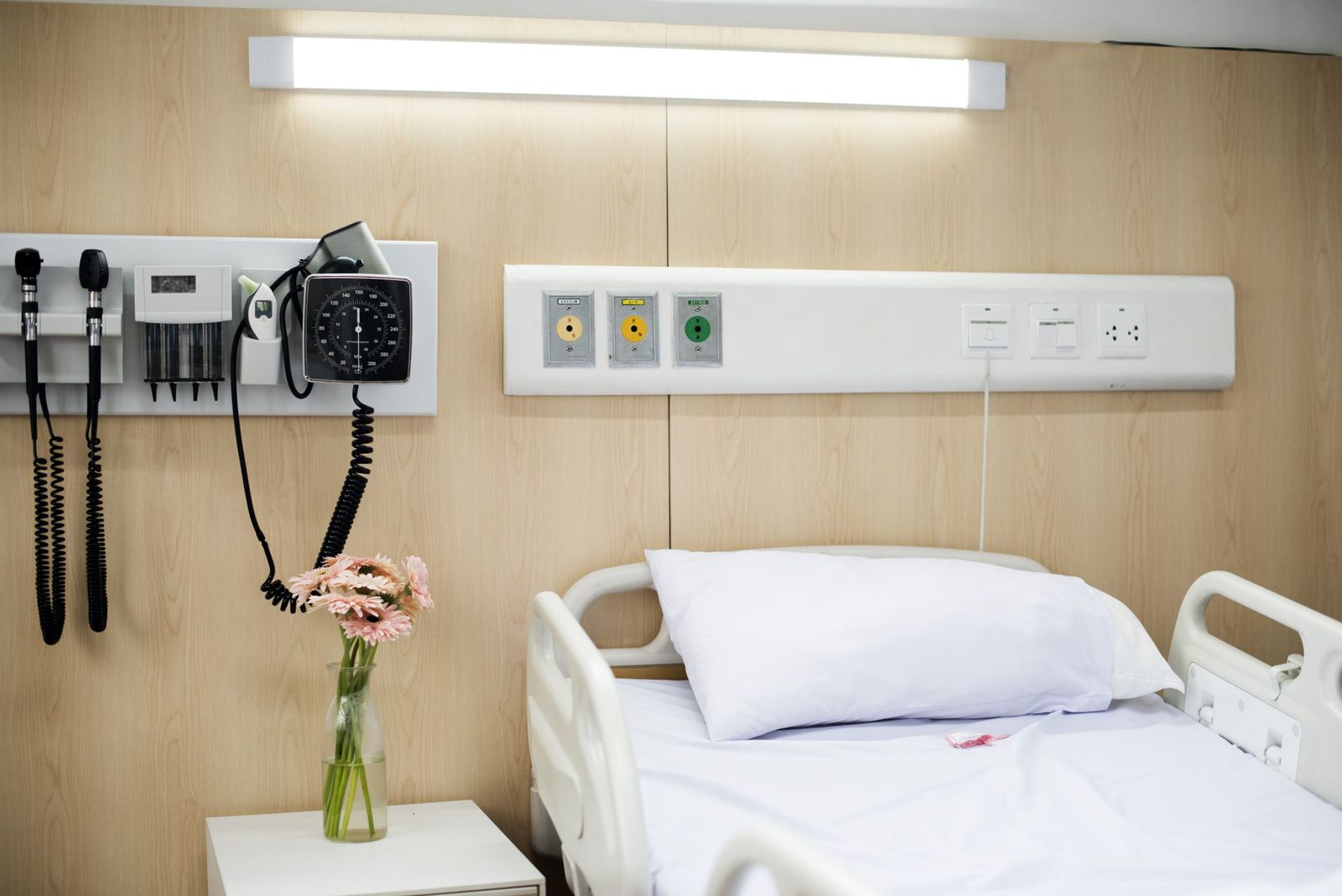 Clean, unoccupied clinical room with a made bed, wall-mounted diagnostic equipment and a vase of pink flowers